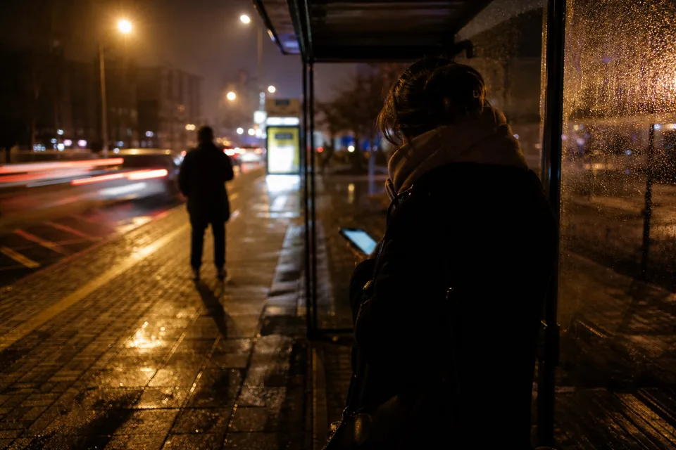 Dorosła kobieta w płaszczu na przystanku nocą, patrzy w telefon, twarz w cieniu